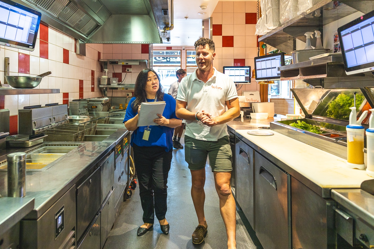 Two restaurant staff walk and talk in a busy stainless-steel kitchen; the woman holds a clipboard and wears a blue top.