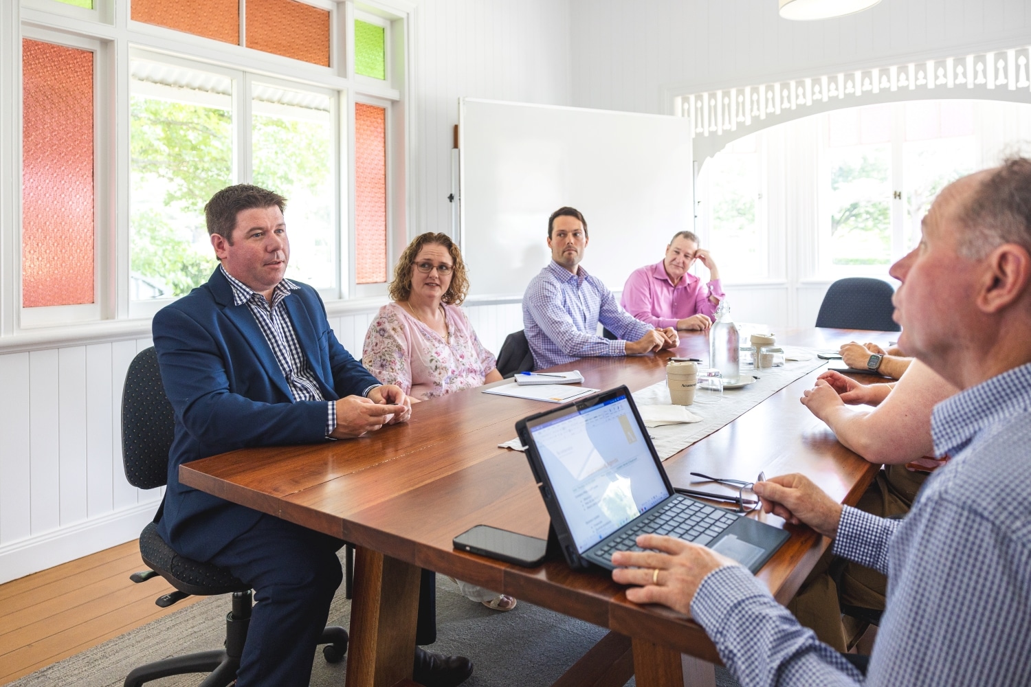 Six professionals sit around a wooden conference table in a bright meeting room, with a laptop and notebooks visible.