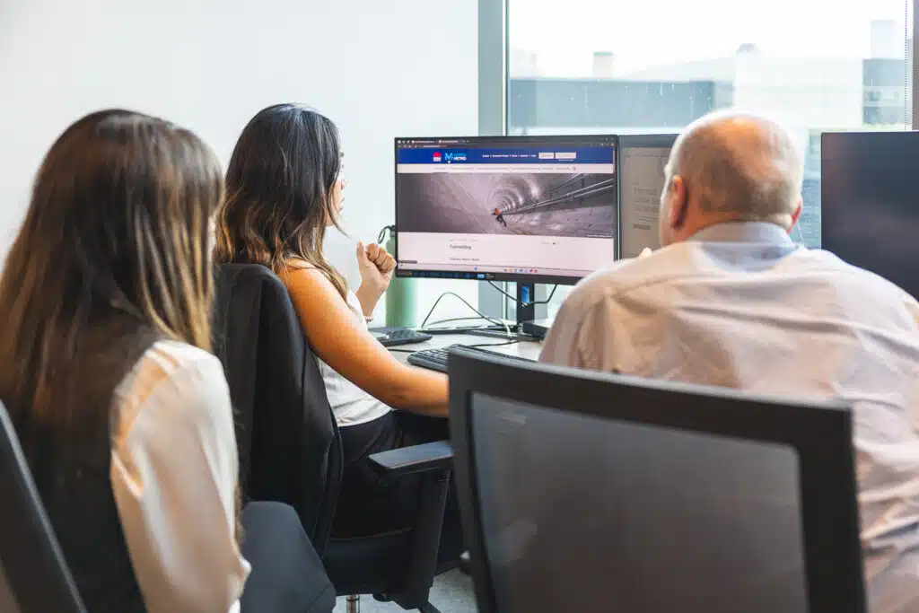 Three professionals at a shared desk looking at a monitor displaying a tunnel image on a webpage.
