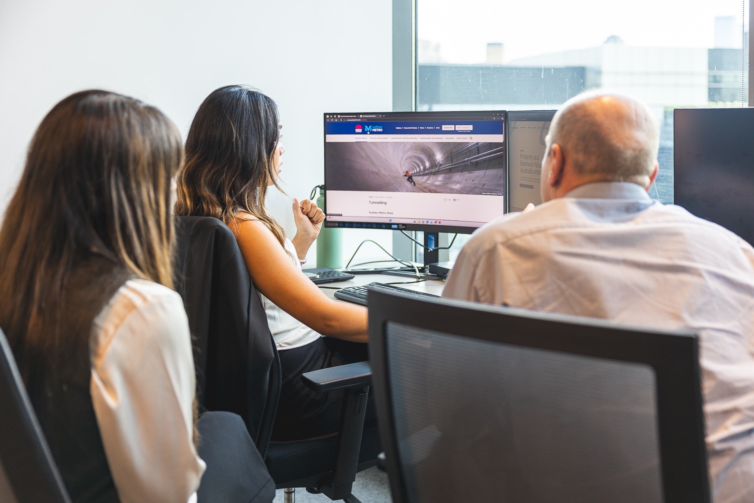 Three professionals at a shared desk looking at a monitor displaying a tunnel image on a webpage.