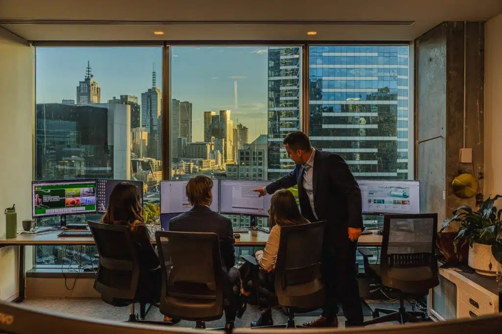 Four coworkers sit at a long desk with multiple monitors while a man in a suit points to a screen, city skyline visible through large windows behind them.