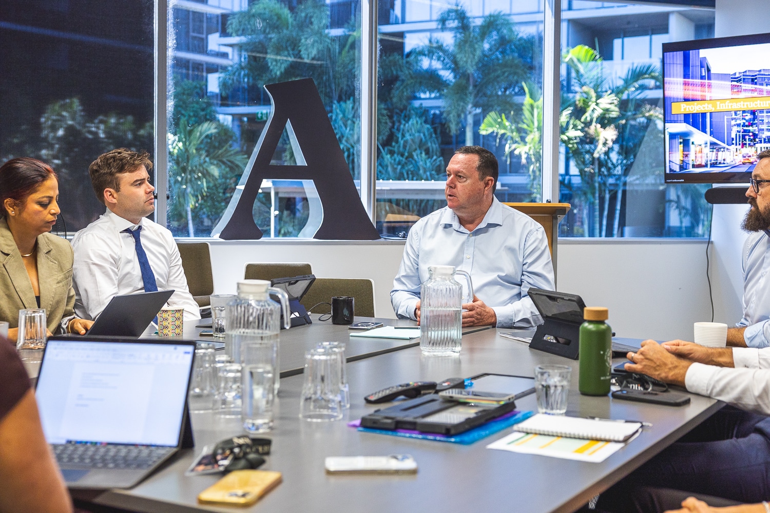 People in a boardroom engaged in a meeting, laptops and notebooks on the table, with a large decorative 'A' sculpture and glass windows behind them.