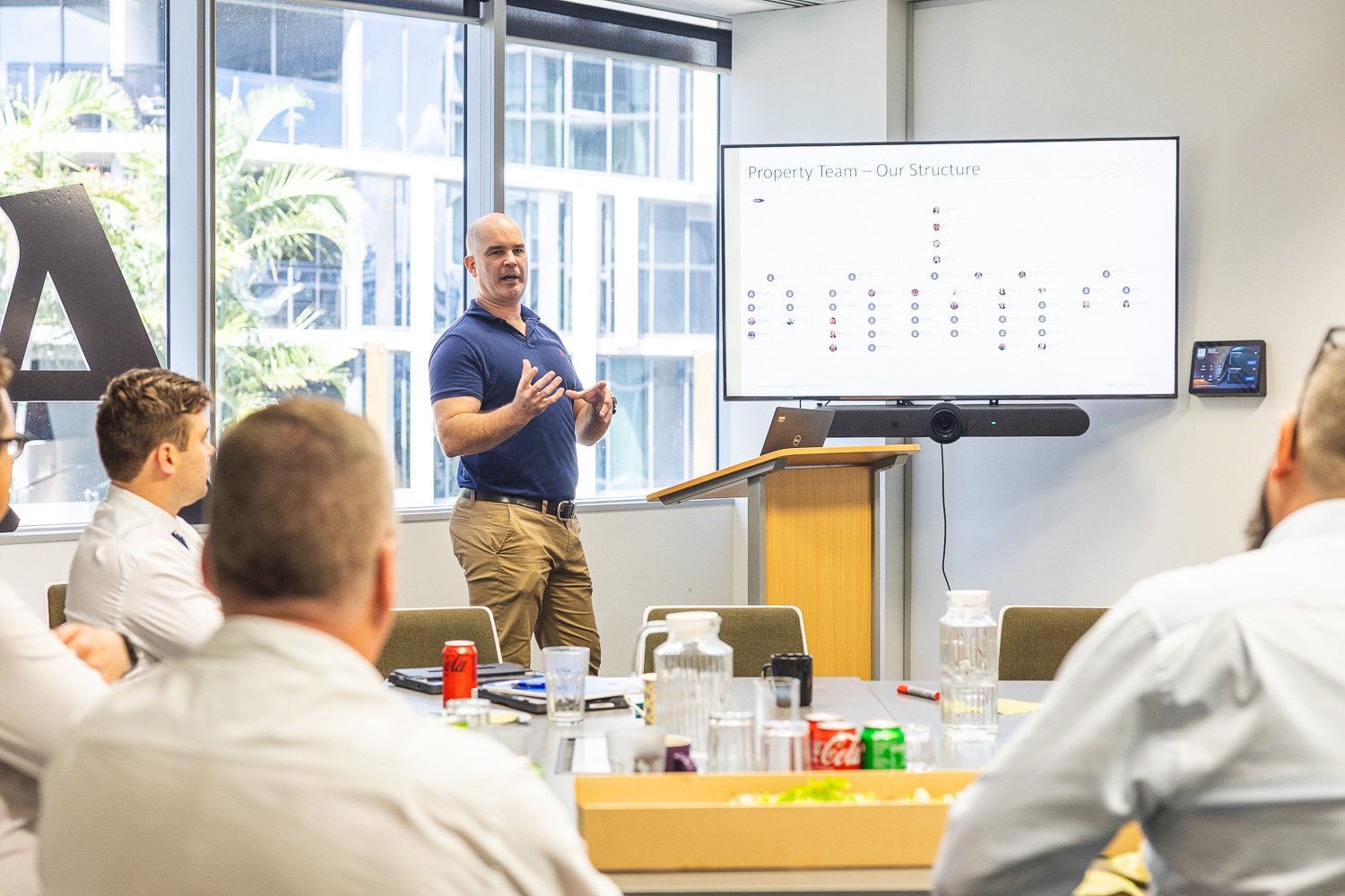 Presenter in a blue polo explains slides to a seated team in a bright conference room with a large screen showing a charted slide.