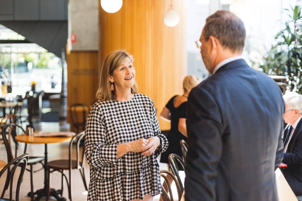 Woman in a black-and-white checkered dress smiles while talking to a man in a suit in a bright cafe setting.