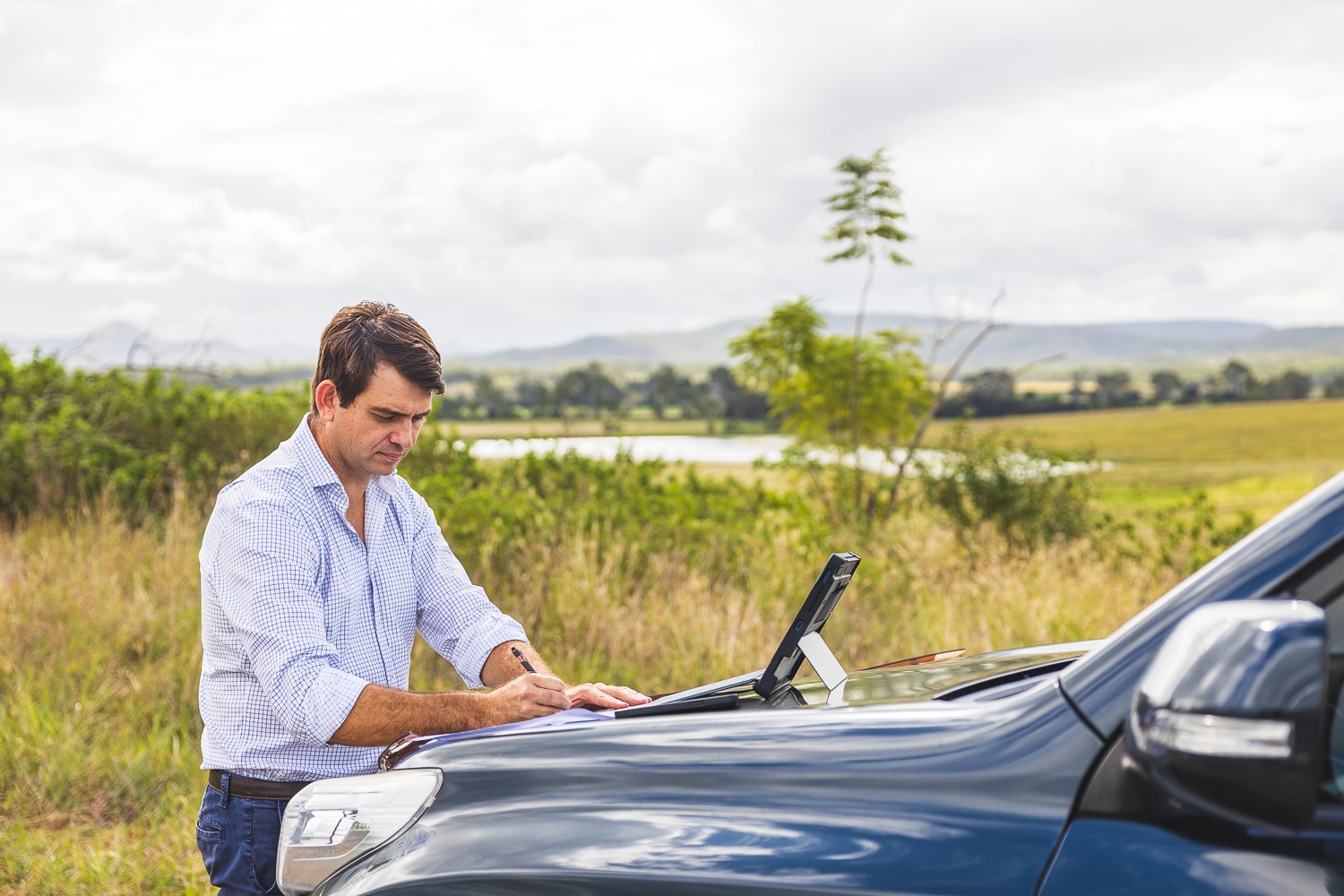 Man in a light checkered shirt writes on paper placed on the hood of a blue vehicle in a rural field.
