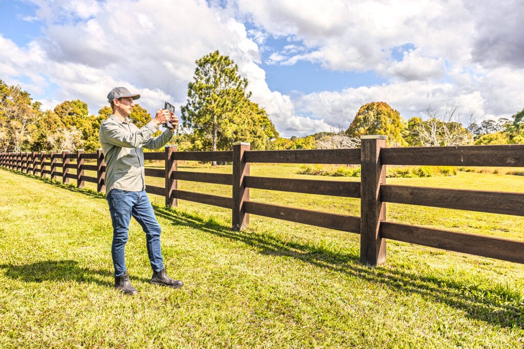 Man in a cap and gray shirt takes a photo with a smartphone along a long wooden fence in a sunny field.