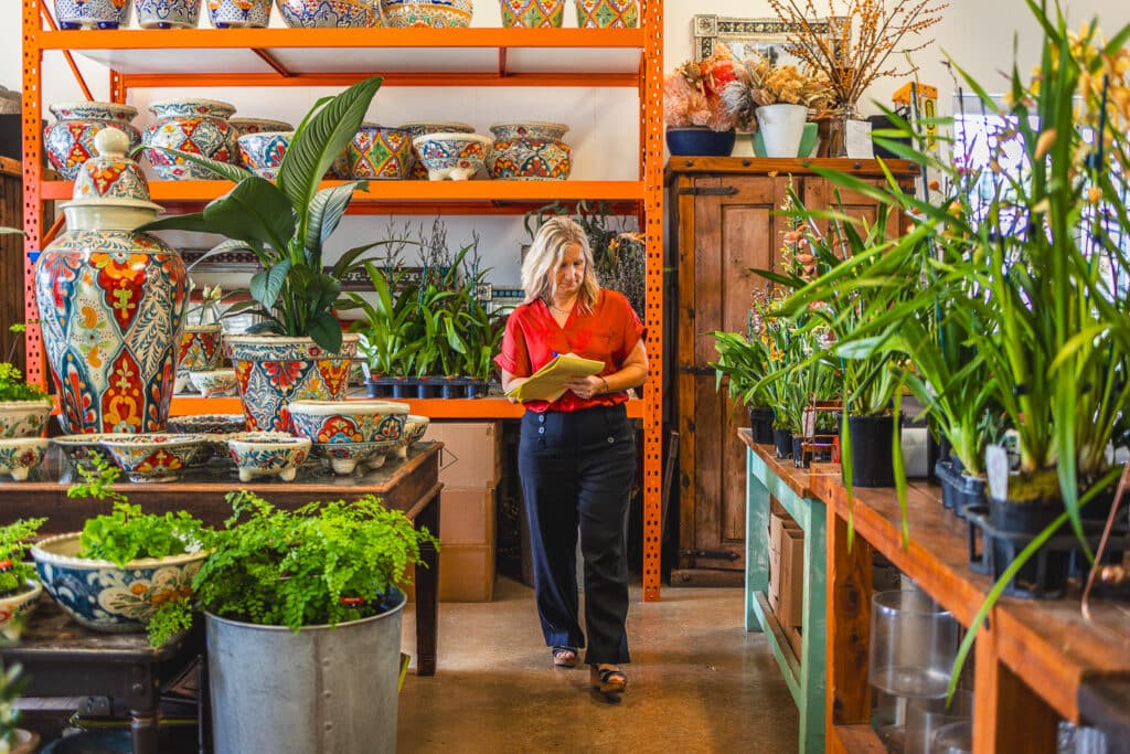 Woman in a red blouse walks through a pottery and plant shop, holding a yellow notebook.
