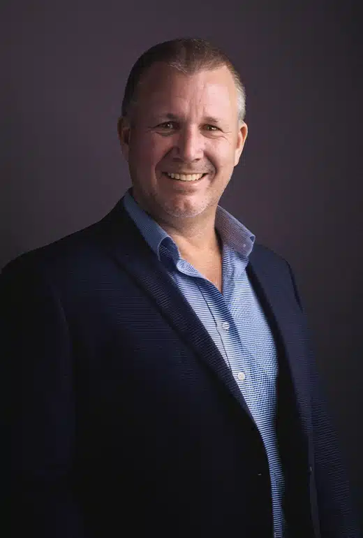 Smiling man in a navy blazer and light blue shirt, standing against a dark backdrop. Used as a professional headshot.