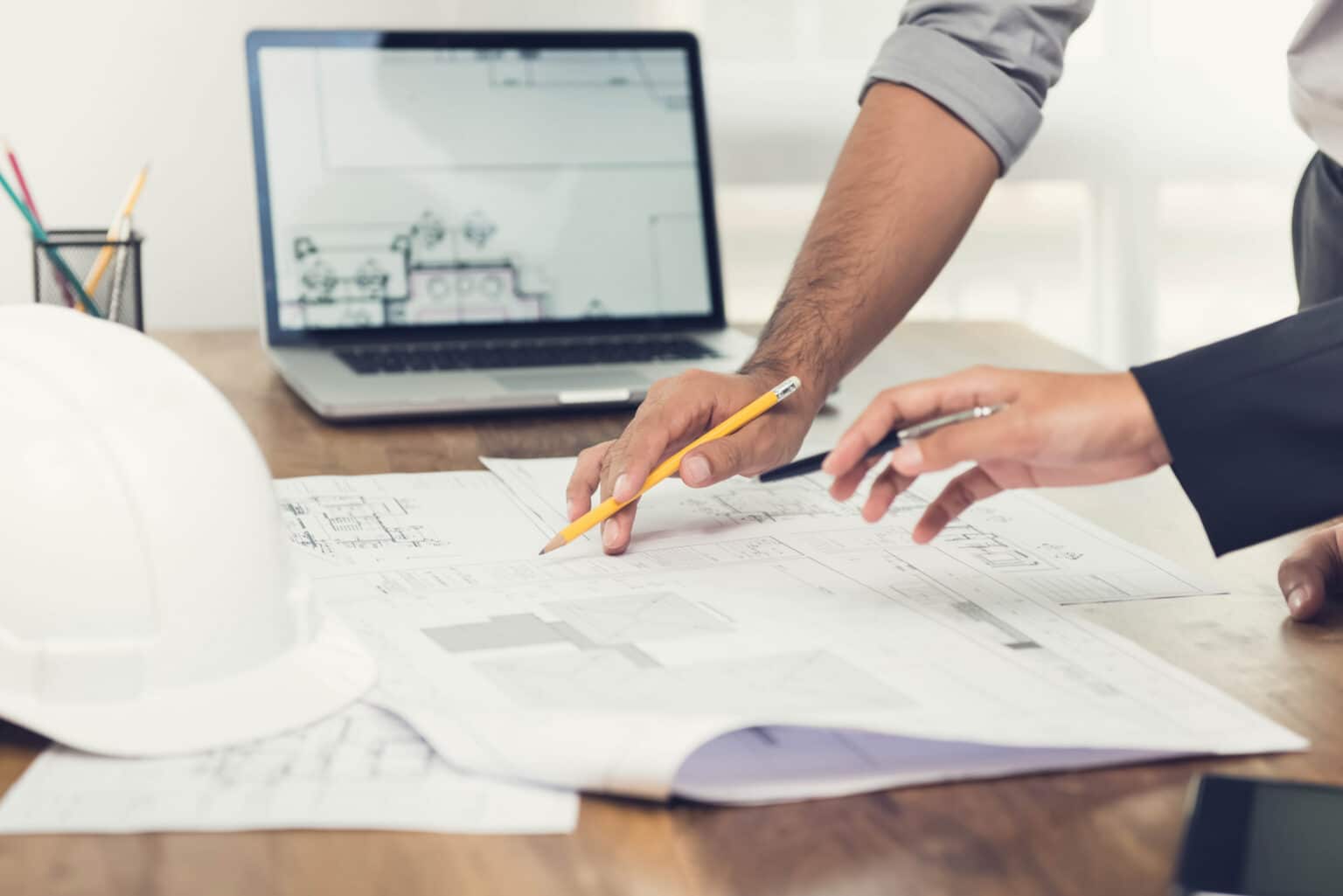Two people reviewing architectural blueprints at a desk with a laptop nearby, pencil in hand, collaborative planning scene